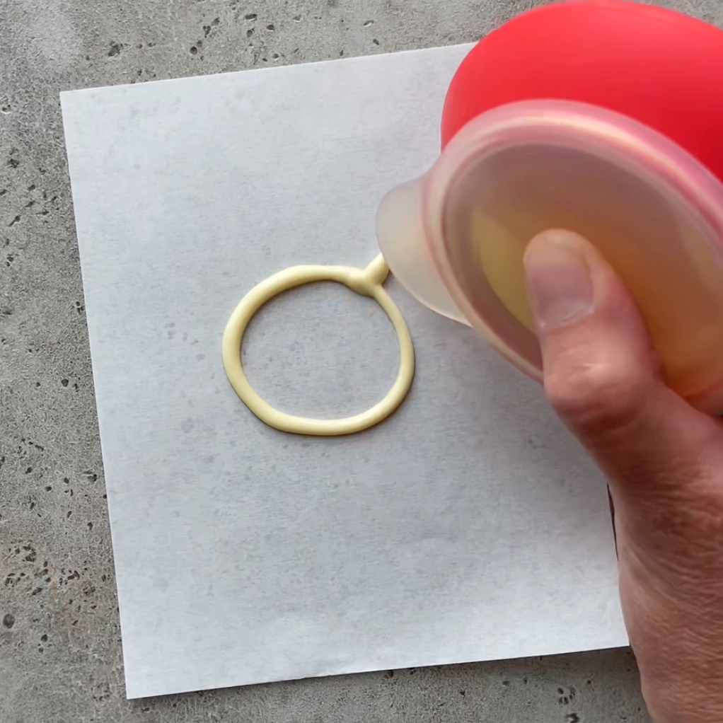 A hand pipes white chocolate from a squeeze bottle onto a square piece of parchment paper, creating a circular shape.
