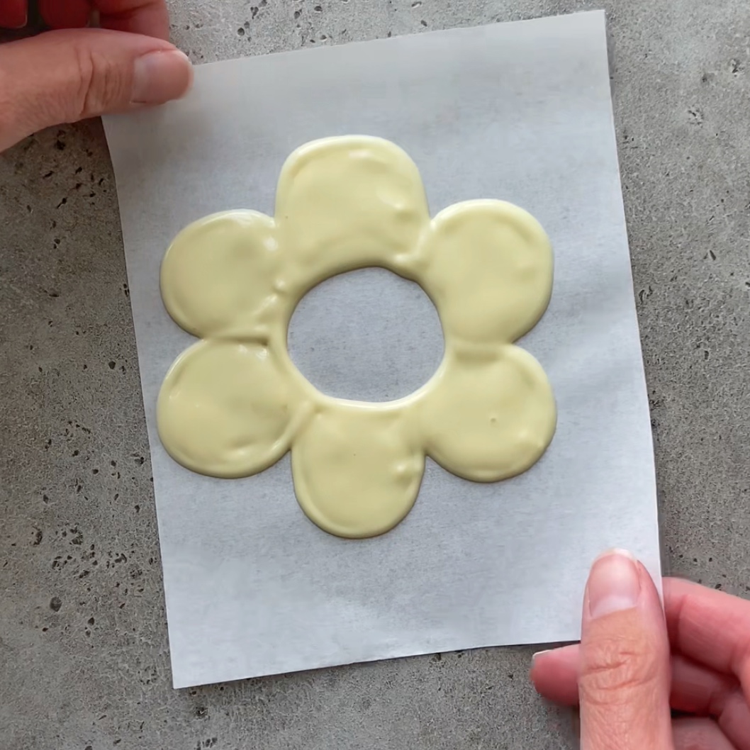 A pair of hands holds a sheet of parchment with white chocolate piped in the shape of a six-petal flower with a hole in the center.