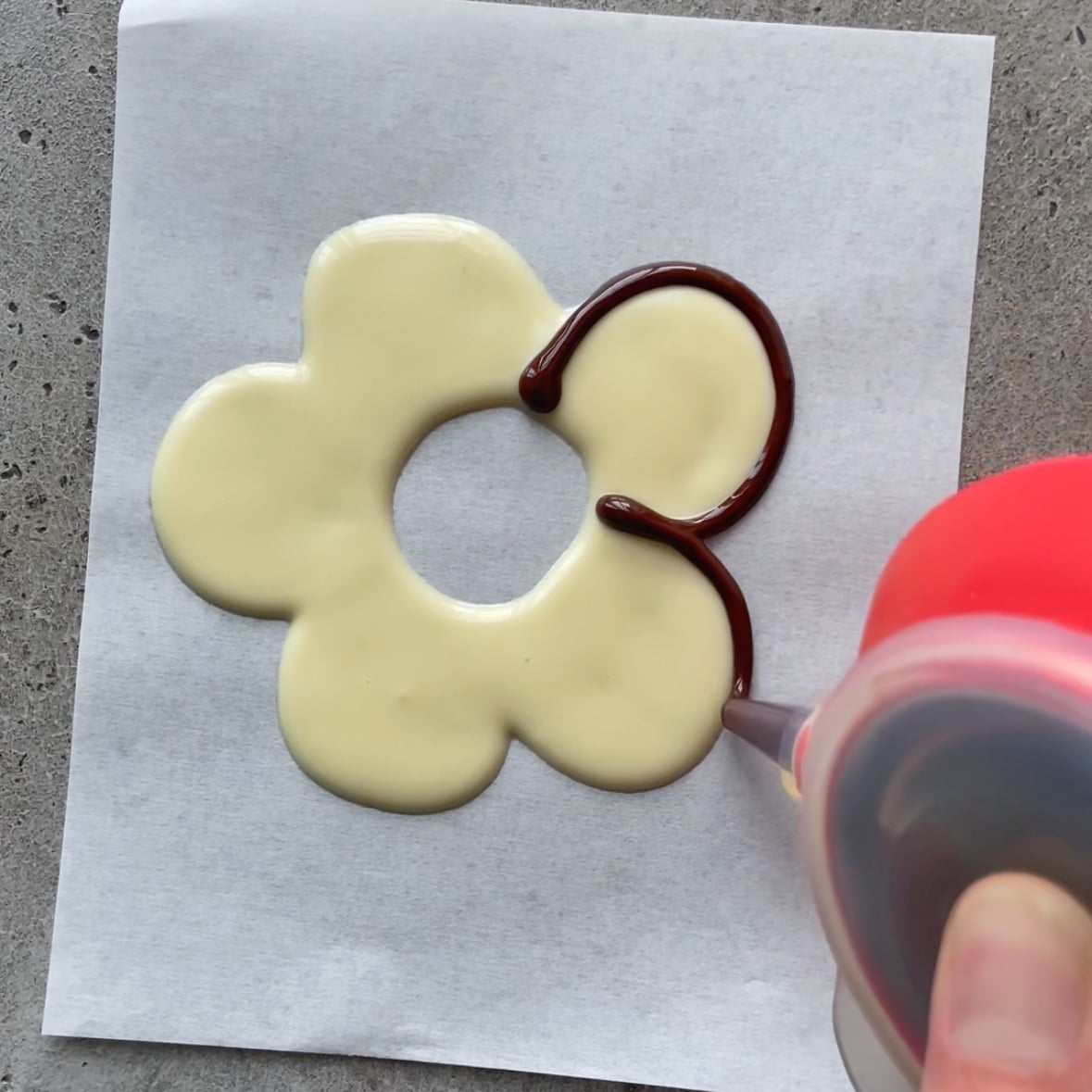 A hand pipes dark chocolate onto the edge of a white chocolate flower shape on parchment paper, all on a gray surface.