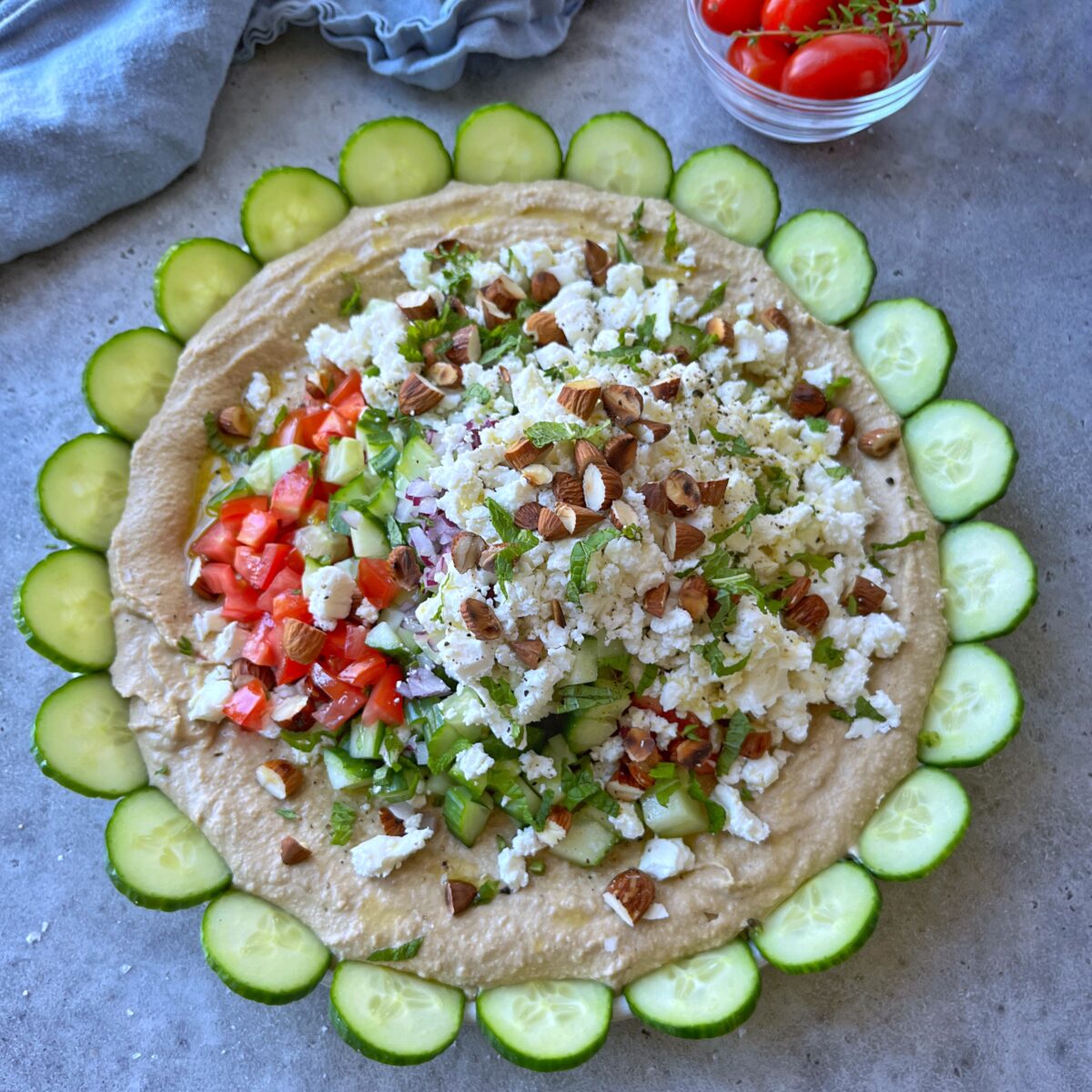 A large layered hummus dip topped with chopped tomatoes, cucumbers, feta cheese, herbs, and almonds, artfully garnished with sliced cucumbers arranged around the edge.