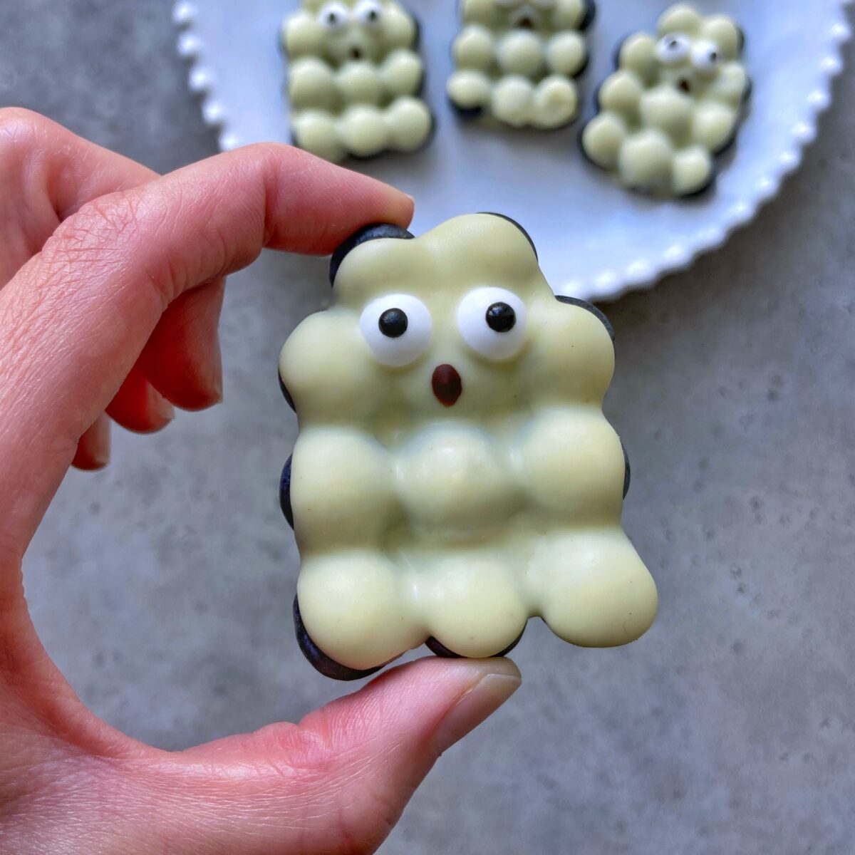 A hand holds a white chocolate blueberry shaped ghost with two candy eyes and a surprised mouth, with more playful treats on a plate in the background.