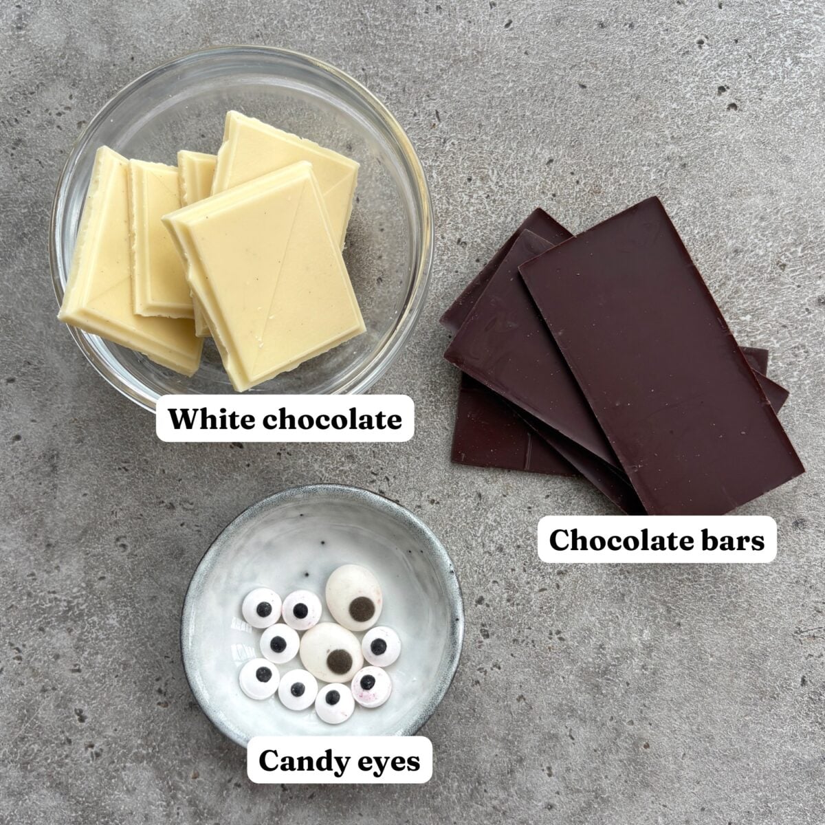 A bowl of white chocolate, a stack of dark chocolate bars, and a bowl of edible candy eyes.