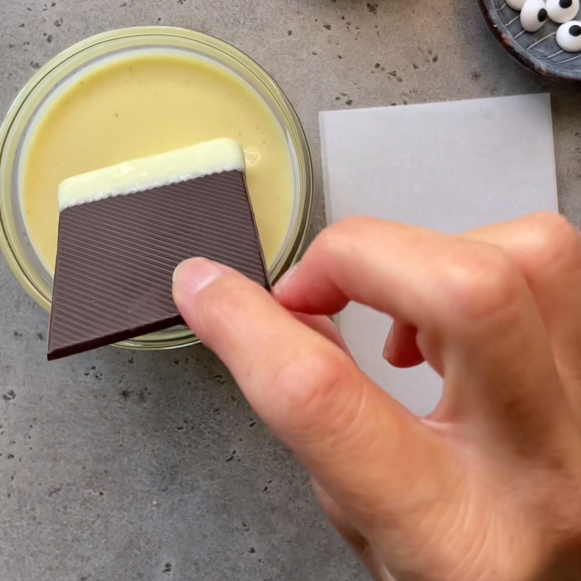 A hand dips a piece of dark chocolate with ridges into a bowl of melted white chocolate on a gray countertop.