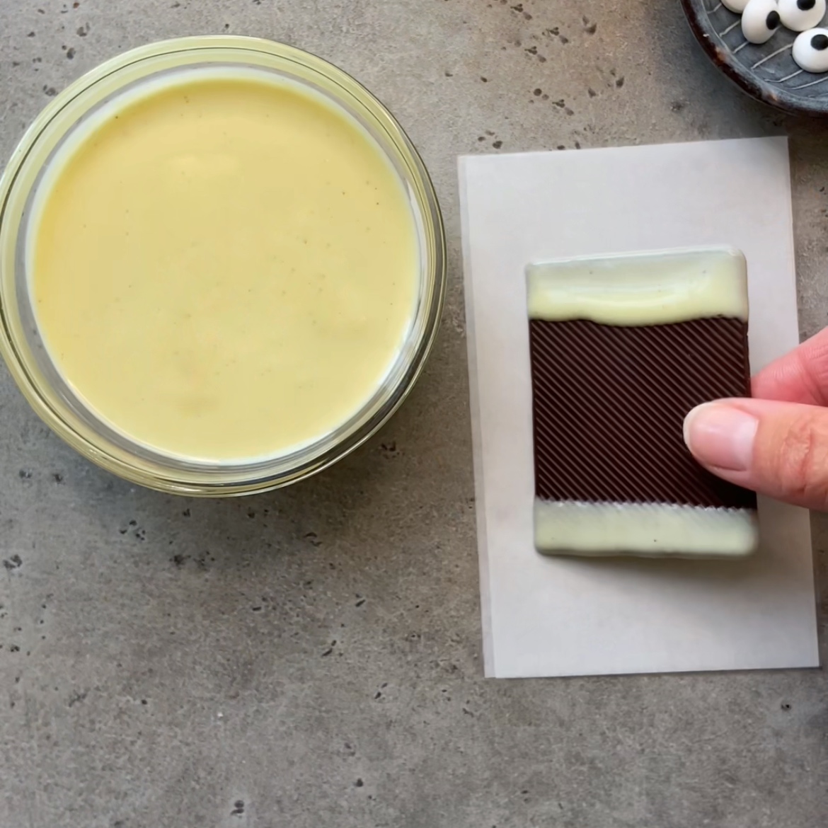 A hand holds a rectangular chocolate partially dipped in white chocolate, next to a bowl of chocolate.