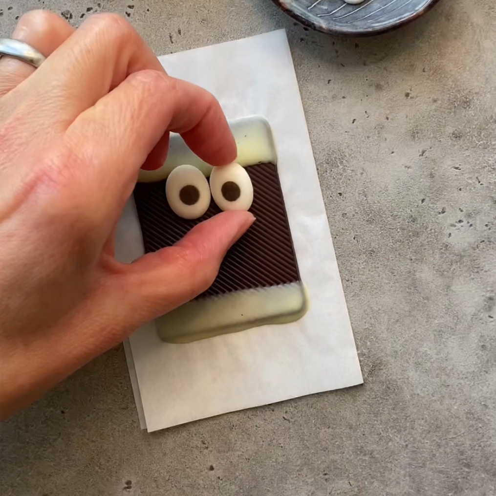 A hand places two candy eyes on a rectangular piece of chocolate with a white chocolate border, resting on parchment paper.