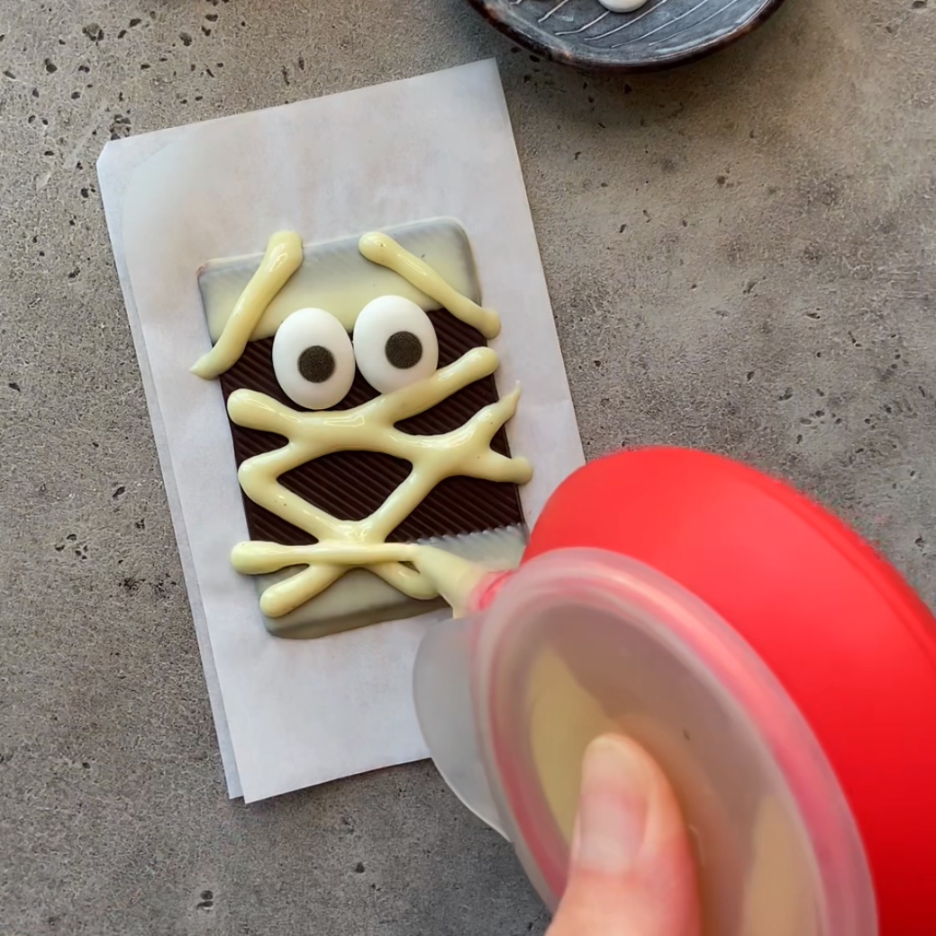 A hand pipes white chocolate in zigzags over a rectangular chocolate bar with candy eyes.