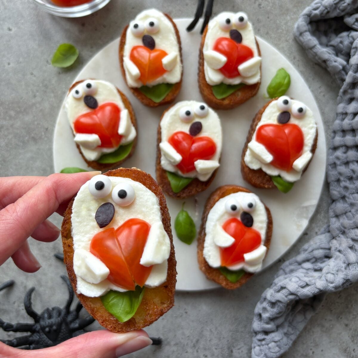 Ghost Toast crostini topped with mozzarella, candy eyes, fig peel mouth, and tomatoes are arranged to look like ghosts holding hearts on a white plate, with a hand holding one piece.