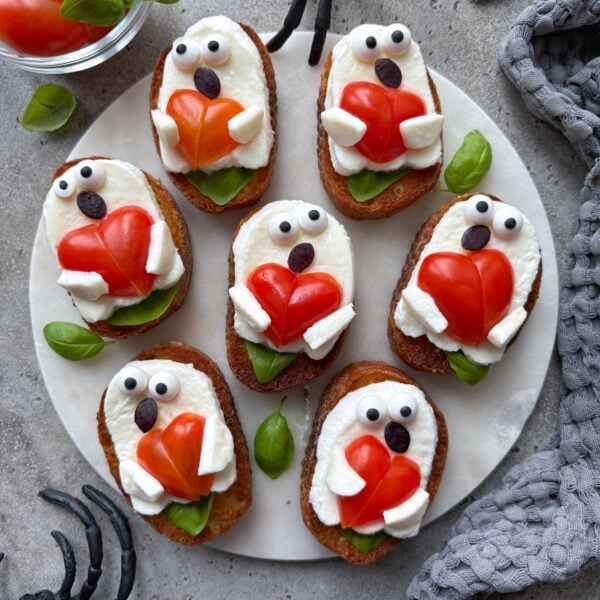 Ghost Toast crostini are topped with mozzarella shaped like playful ghosts holding heart-shaped tomato pieces, with candy eyes and a fig peel mouth on a round platter.
