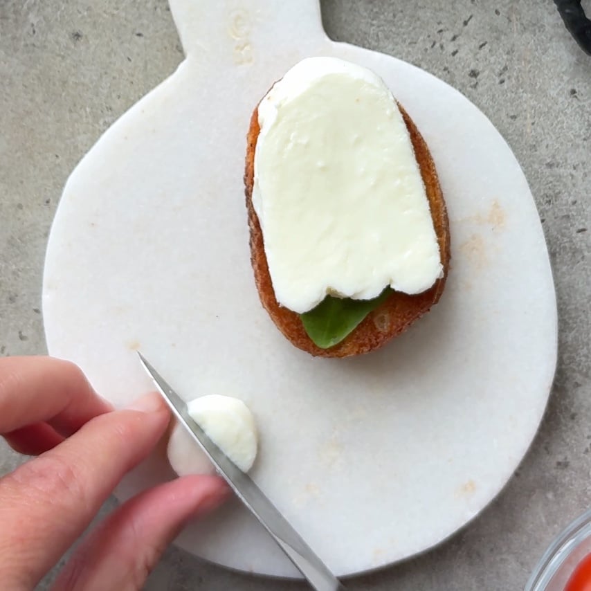 A hand slices mozzarella cheese disc next to a piece of bread topped with mozzarella ghost and a basil leaf.