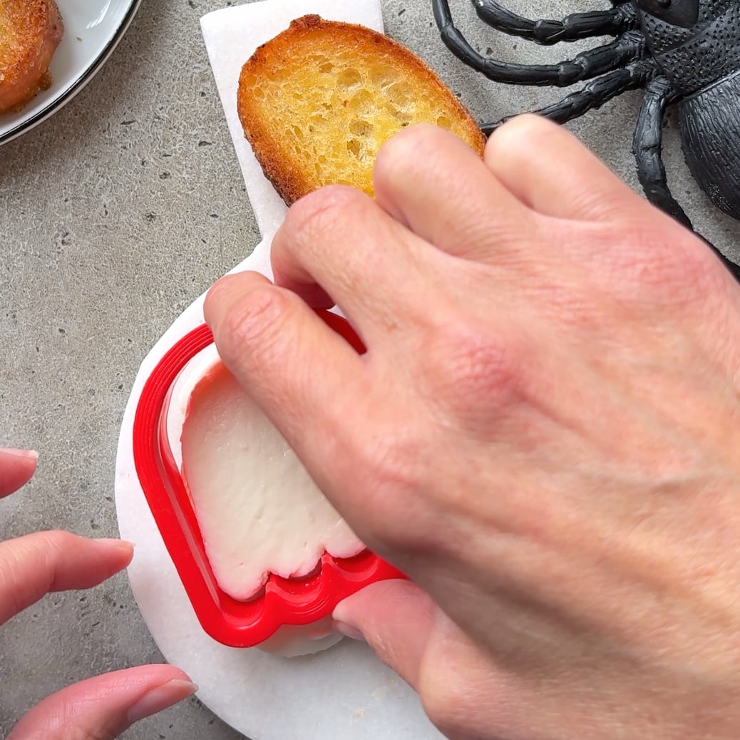 A hand presses a red ghost-shaped cookie cutter into white cheese on a round marble plate, with toast and a plastic spider nearby.