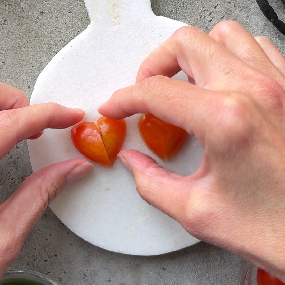 Two hands arrange two halves of a grape tomato into a heart shape on a white marble cutting board.