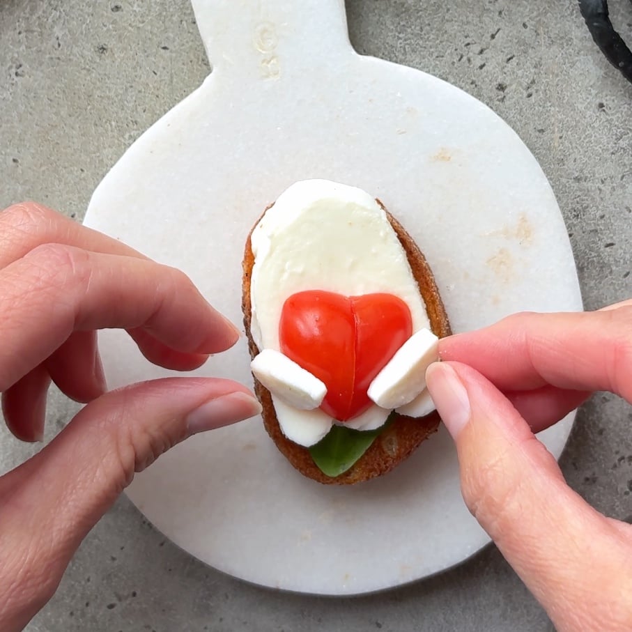 Hands arranging cheese slices on a piece of bread topped with a basil leaf, a mozzarella ghost, and a halved grape tomato shaped like a heart.