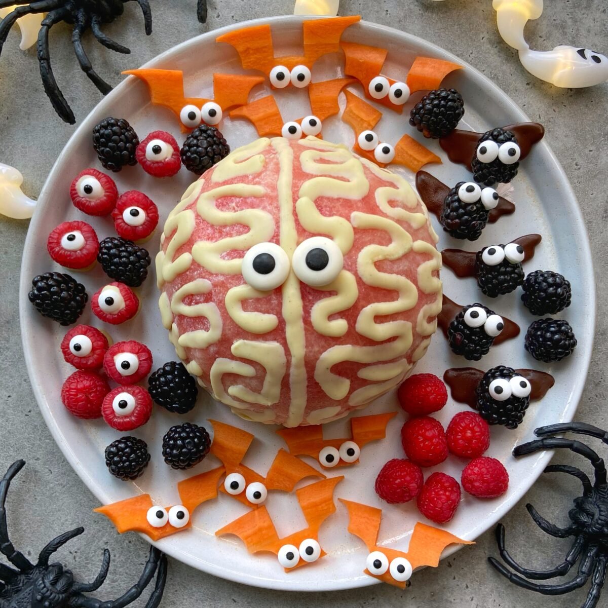 A halved watermelon decorated to look like a brain is surrounded by fruit and vegetable bites shaped as bats, spiders, and monsters with candy eyes on a white plate.