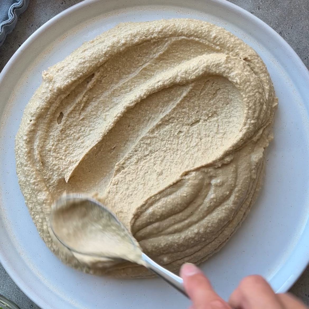 A hand uses a spoon to spread creamy beige layered hummus dip in a circular pattern on a white plate.