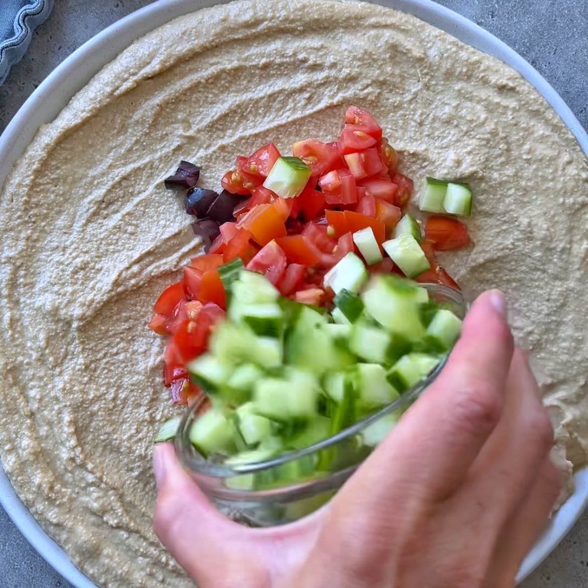 A hand adds diced cucumber to a plate, creating a vibrant layered hummus dip with chopped tomatoes and olives.