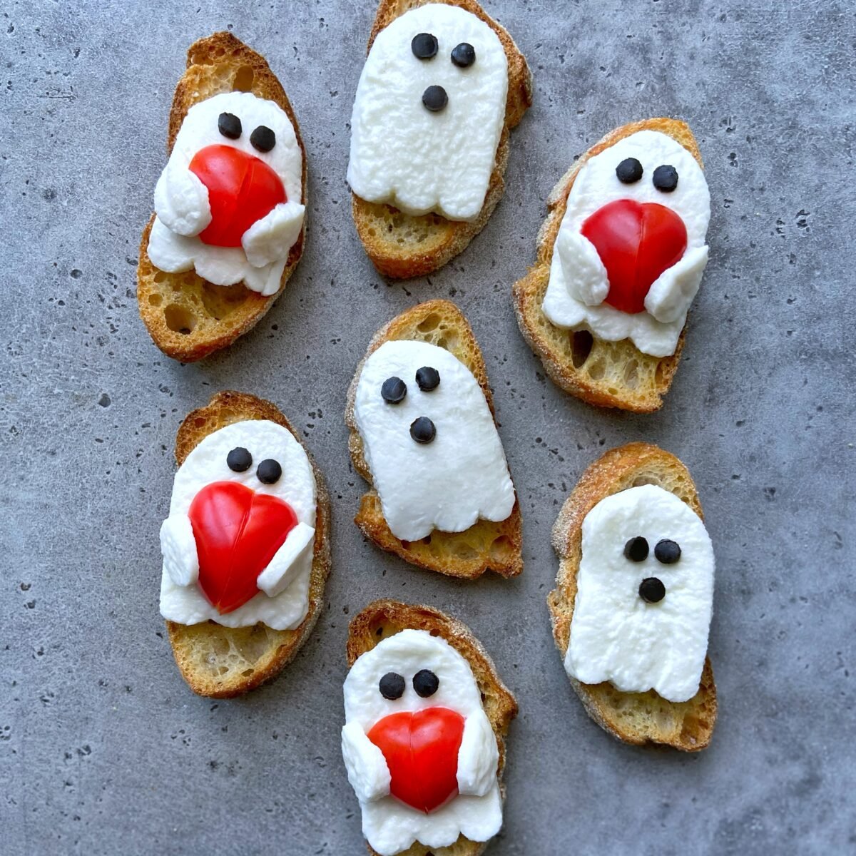 Seven pieces of bread are topped with cheese arranged to look like ghosts. All have small black olive circles for eyes, and some are holding red heart-shaped tomatoes, all on a gray surface.