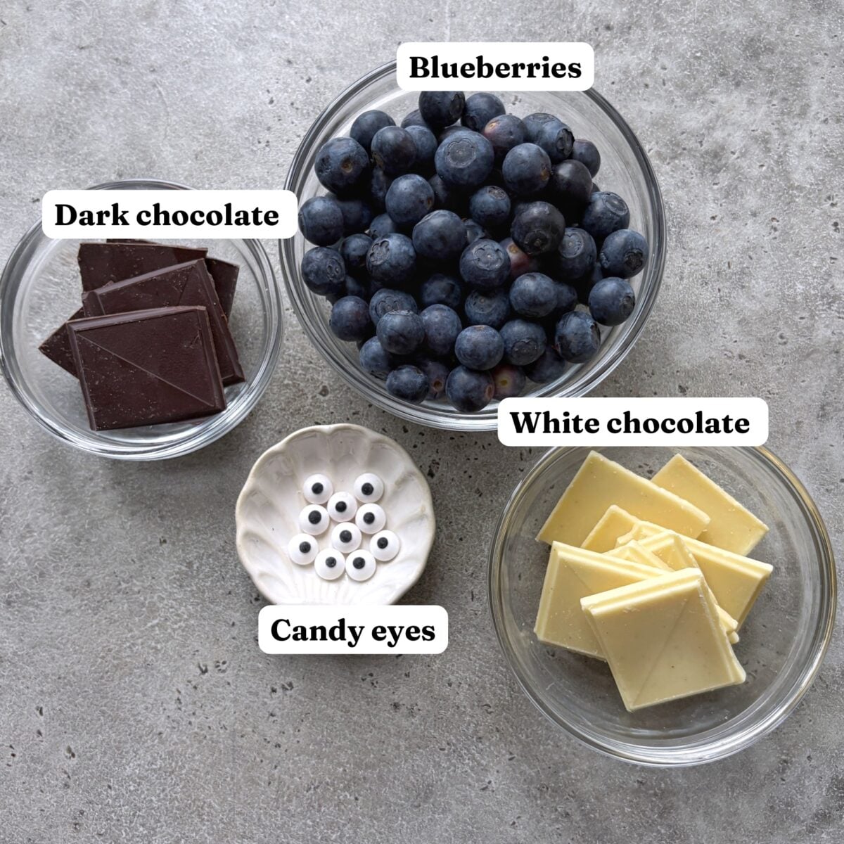 Four small bowls containing dark chocolate, blueberries, white chocolate, and candy eyes.