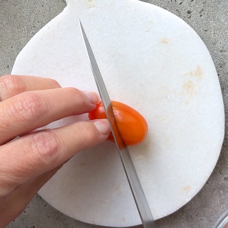 A hand slices a small orange tomato at a slant with a knife on a round white cutting board.