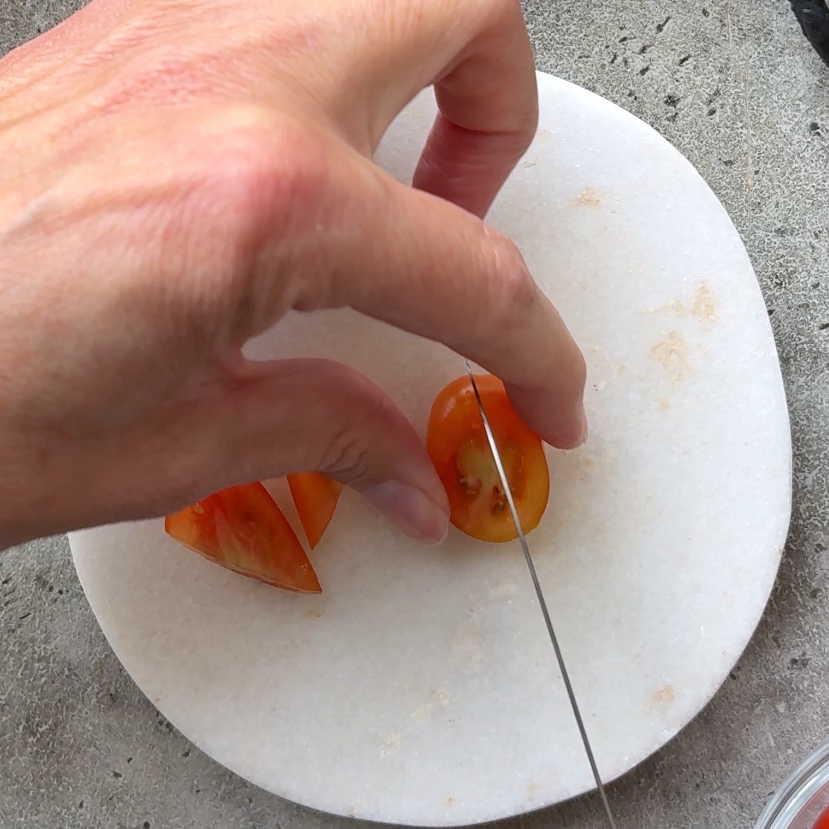 A hand holds a halved grape tomato on a white cutting board, slicing it in half with a knife.