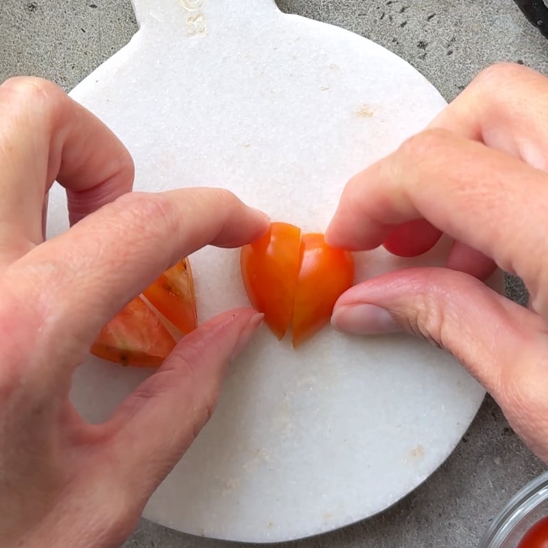 Two hands turn two tomato halves to face each other, making a heart shape on a round white cutting board.