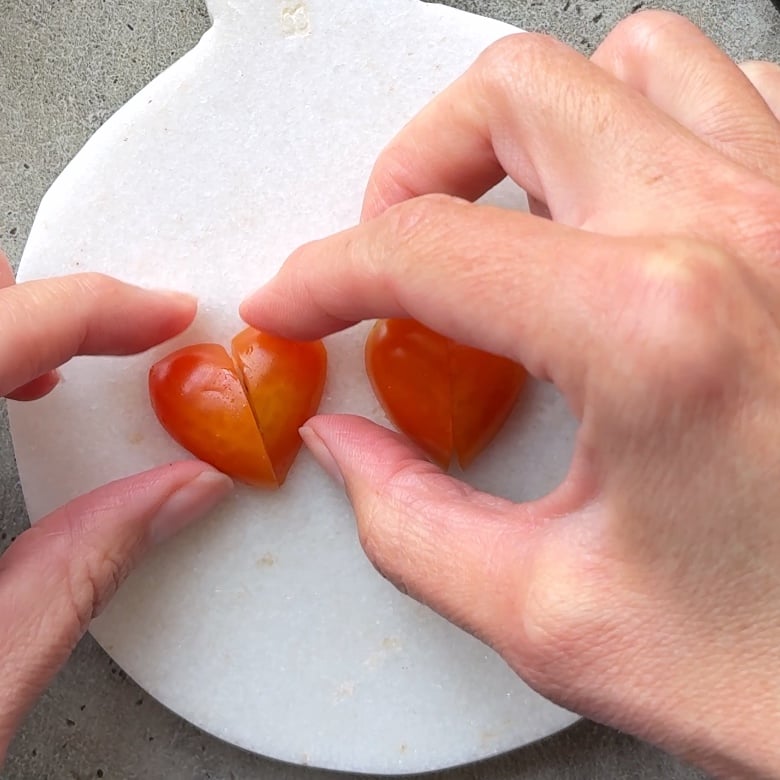 Two hands holding halves of a red grape tomato cut to form a heart shape on a round white cutting board.