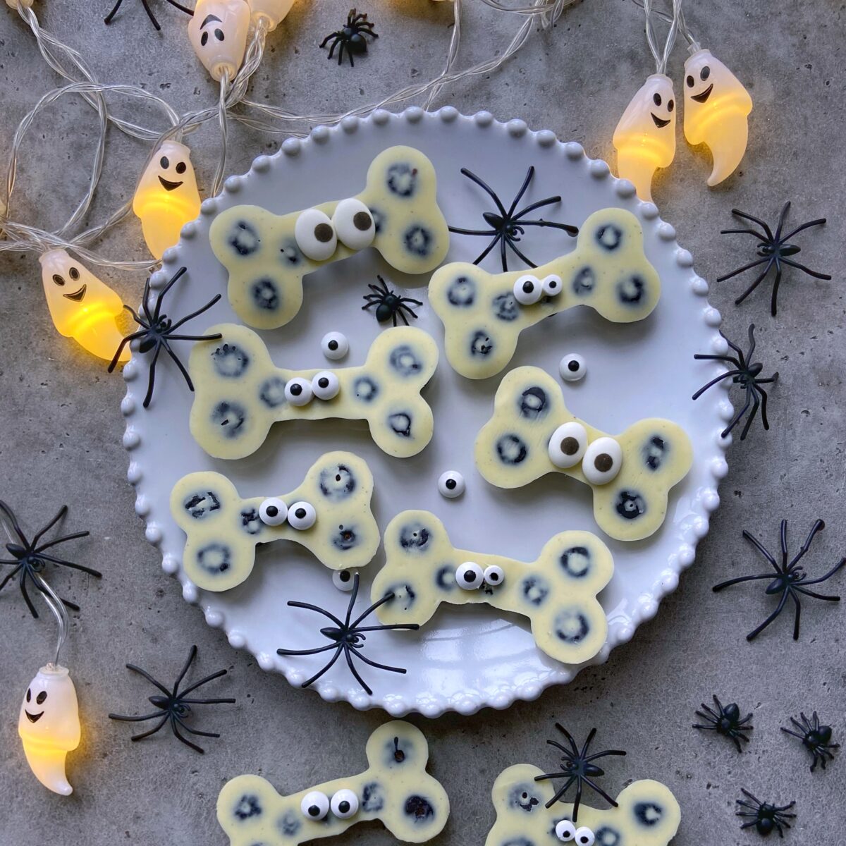 A plate of white chocolate bone-shaped treats with candy eyes, surrounded by plastic spiders and ghost string lights on a gray surface.