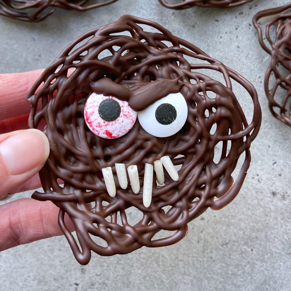 A hand holds a round treat with candy eyes, white candy teeth, and a tangled appearance.
