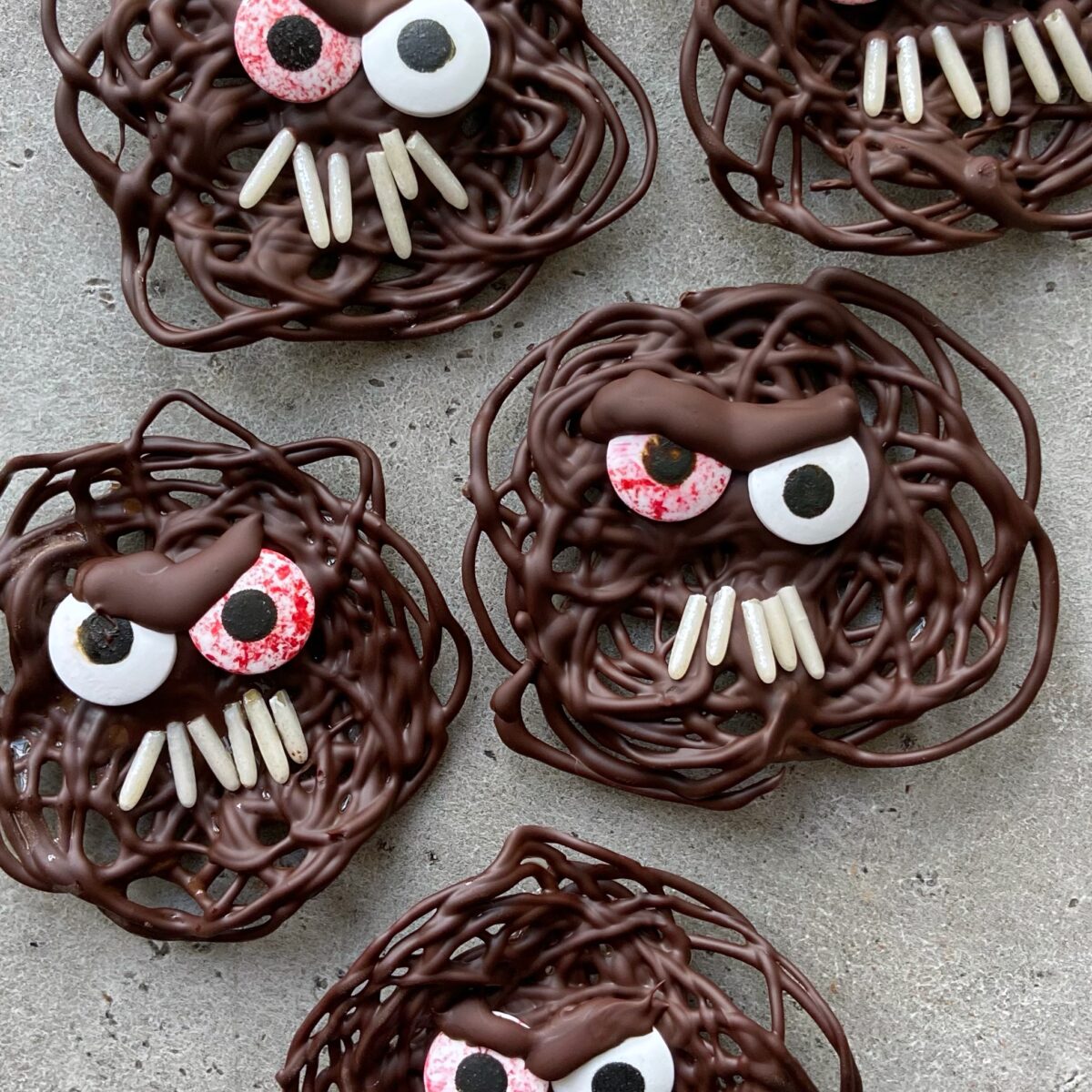 Five Chocolate Monsters shaped like monster faces, complete with candy eyes and sprinkle teeth, are arranged on a gray surface.