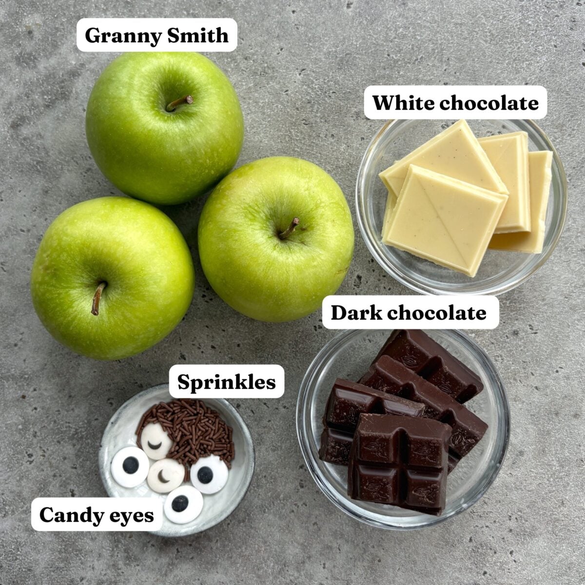 Three Granny Smith apples, bowls of white and dark chocolate, and a small dish of sprinkles and candy eyes.