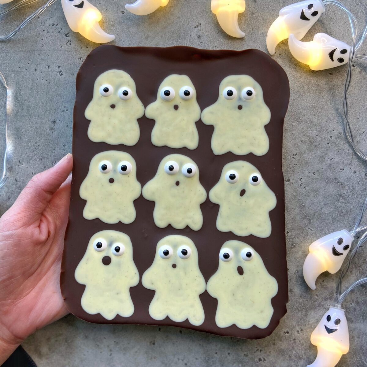 A hand holds a chocolate slab topped with nine white chocolate ghost bites, each with candy eyes, surrounded by ghost-shaped string lights on a gray surface.