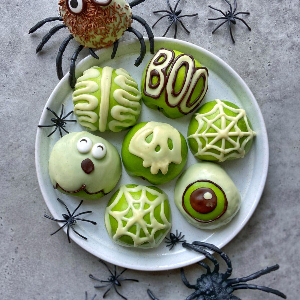 A white plate with seven decorated green and white Halloween-themed bites, including spider, brain, skull, eyeball, web, and “BOO” designs, surrounded by plastic spiders.