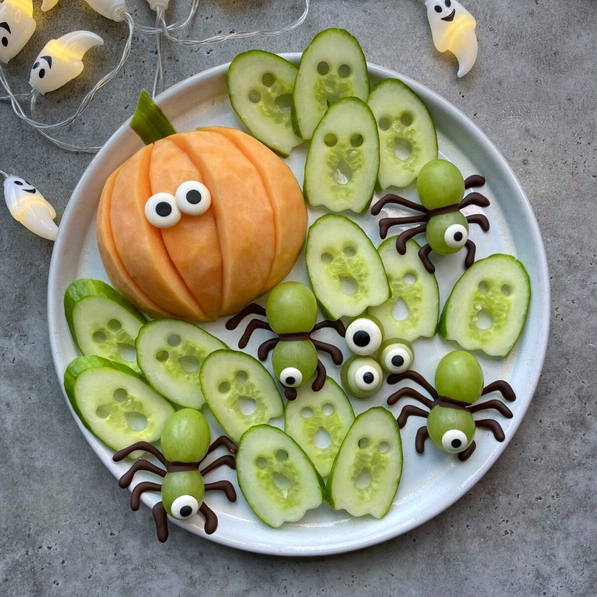 A plate with cucumber slices, a melon pumpkin with candy eyes, and grape spiders with pretzel legs, arranged as fun Halloween bites.