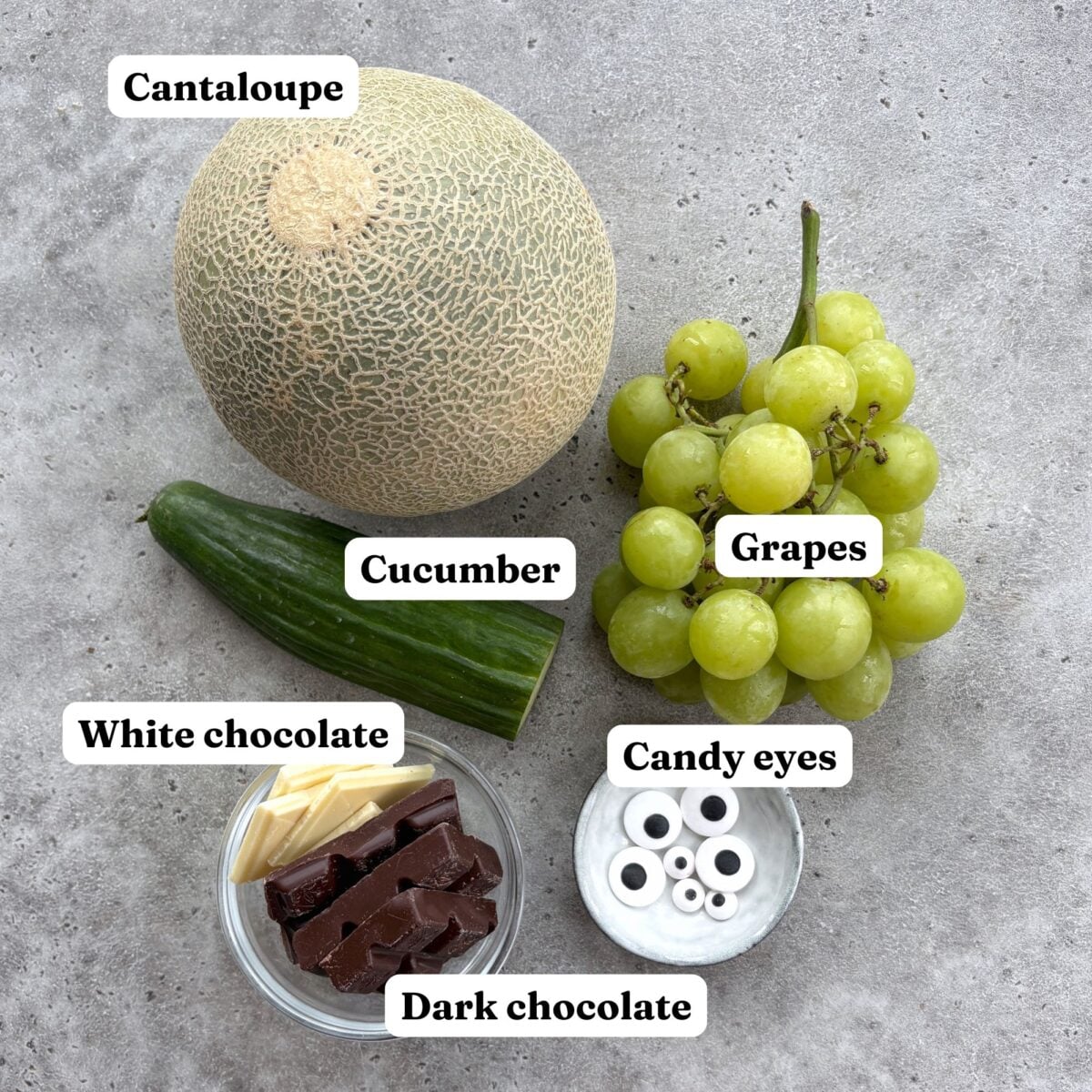 A cantaloupe, a cucumber, a bunch of green grapes, white and dark chocolate pieces, and candy eyes.