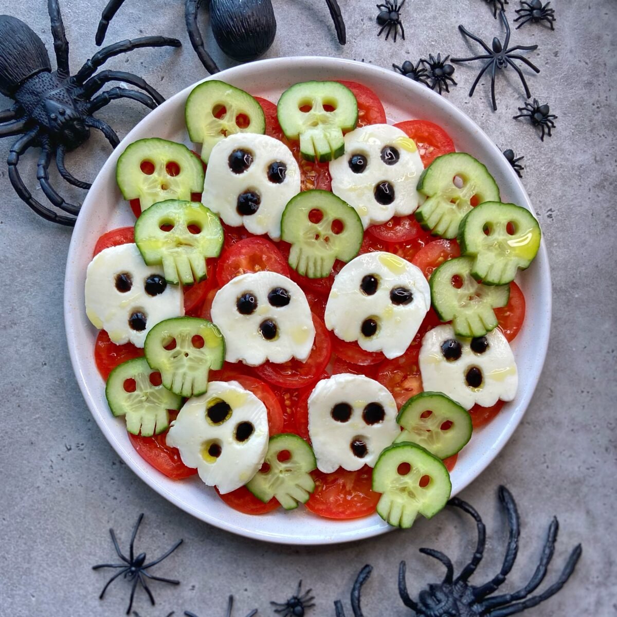 A creative salad with tomato slices, mozzarella ghosts, and cucumber bites shaped like skulls, garnished with balsamic glaze, surrounded by plastic spiders on a gray surface.