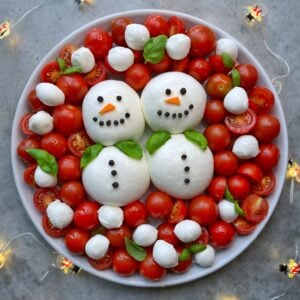 Two snowmen made out of burrata, peppercorns, carrot, and fresh basil are arranged on a platter and surrounded by cherry tomatoes and bocconcini snow.