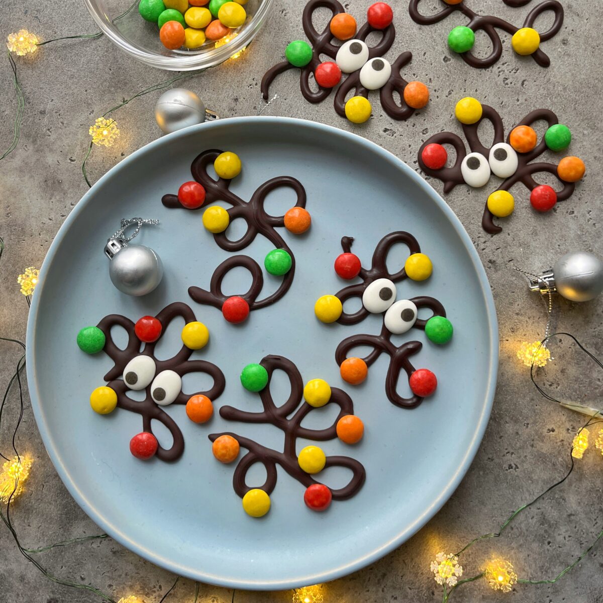 Chocolate shapes decorated with candy eyes and colorful round candies are arranged on a blue plate and a gray surface, surrounded by ornaments and string lights.