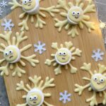 Chocolate Snowflakes-shaped treats with marshmallow centers, decorated with candy eyes and faces, are arranged on a wooden board.