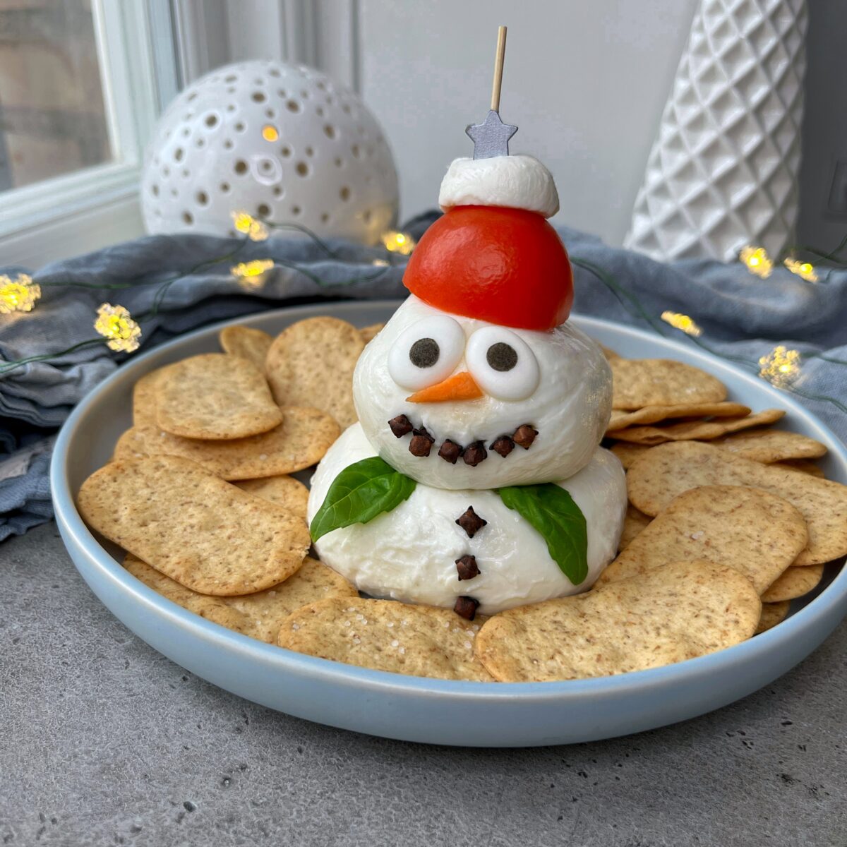 A snowman-shaped cheese ball decorated with a tomato hat and cloves, surrounded by crackers on a plate.