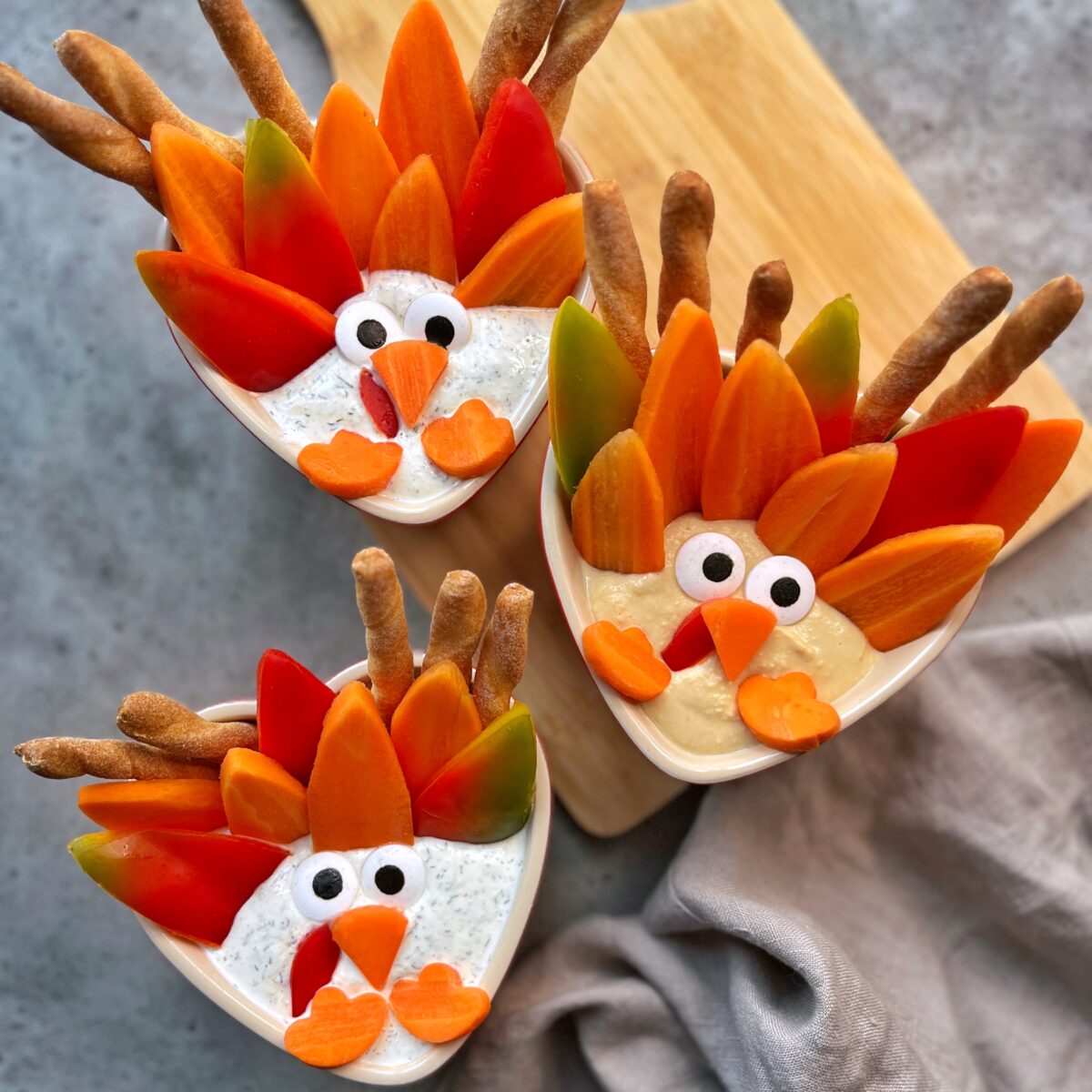 Three bowls of dip are decorated with vegetables and breadsticks arranged to resemble turkeys with carrots, peppers, and candy eyes.