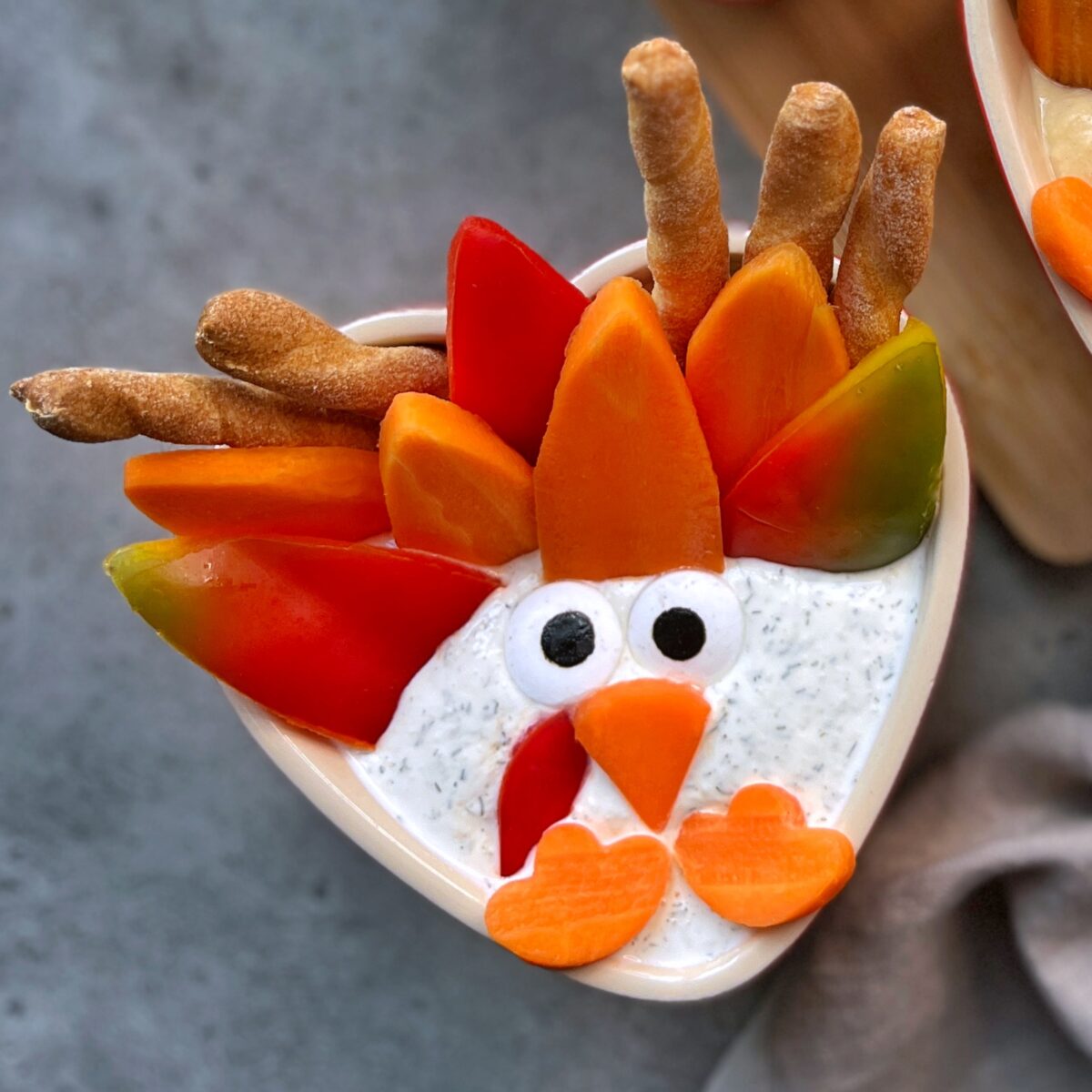 Heart-shaped bowl of dip decorated with vegetables and breadsticks to resemble a turkey using sliced peppers as feathers, carrot pieces for the beak and wattle.