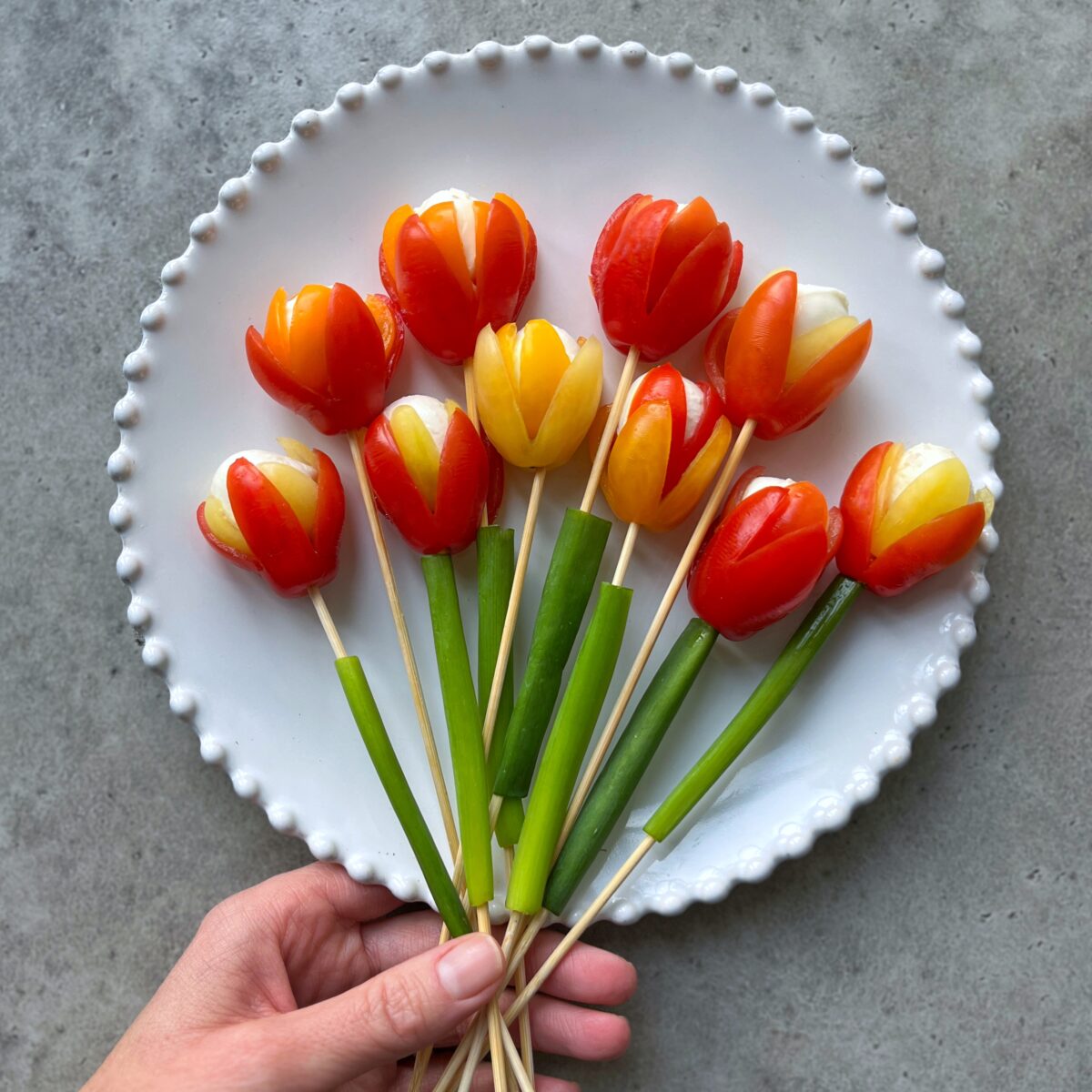 A full view of a hand holding skewered red and yellow tomato flowers with mozzarella and green onions on a white, beaded-edge plate.