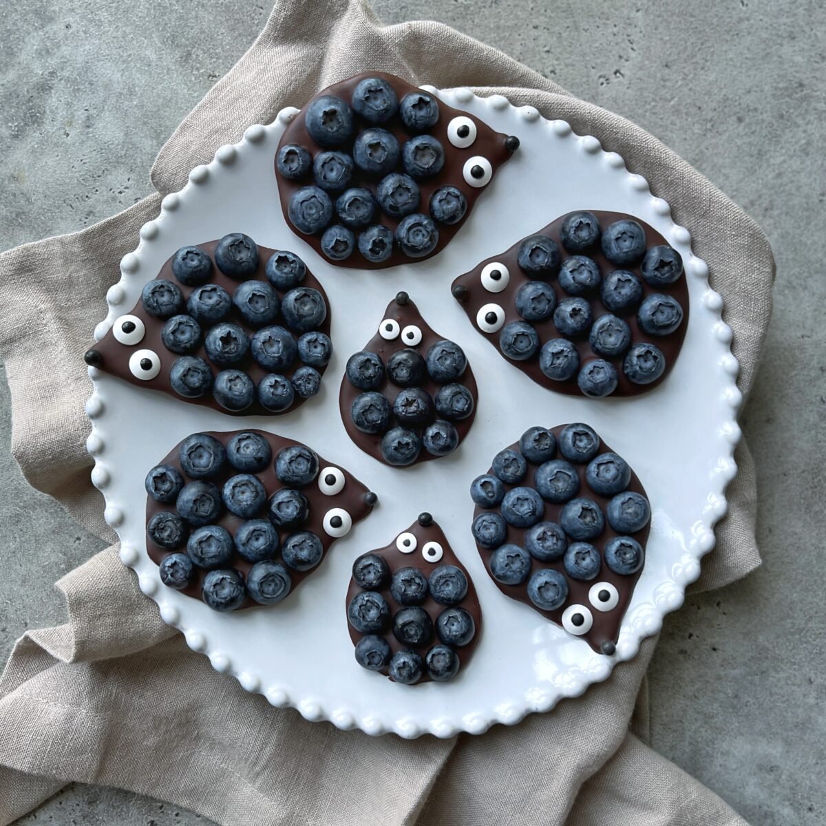 A white plate holds seven No-Bake Chocolate Hedgehog Treats, each shaped like a hedgehog and decorated with blueberries as spikes and candy eyes, placed on a beige cloth.