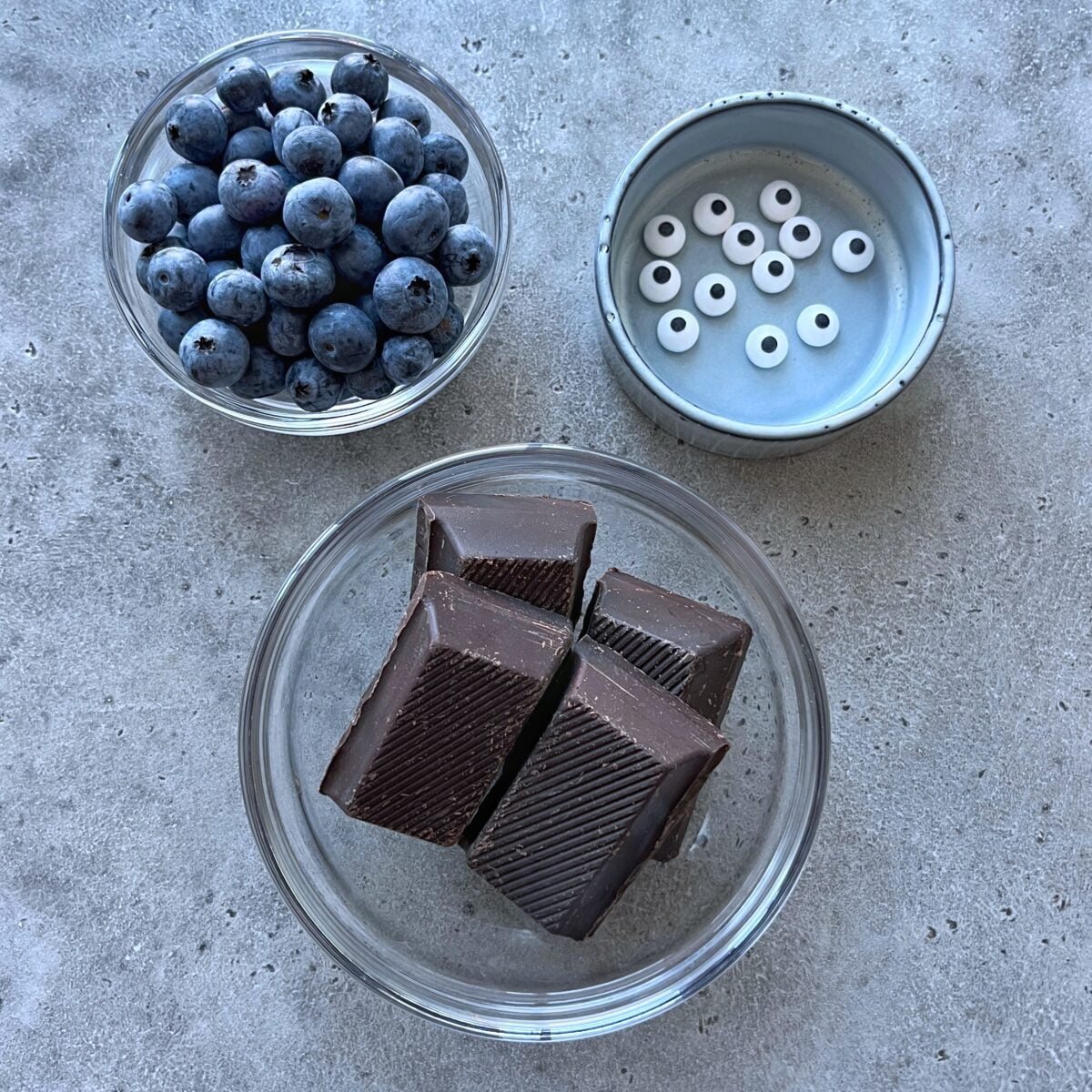Three bowls on a gray surface: one with blueberries, one with chocolate pieces, and one with small round white candy eyes.