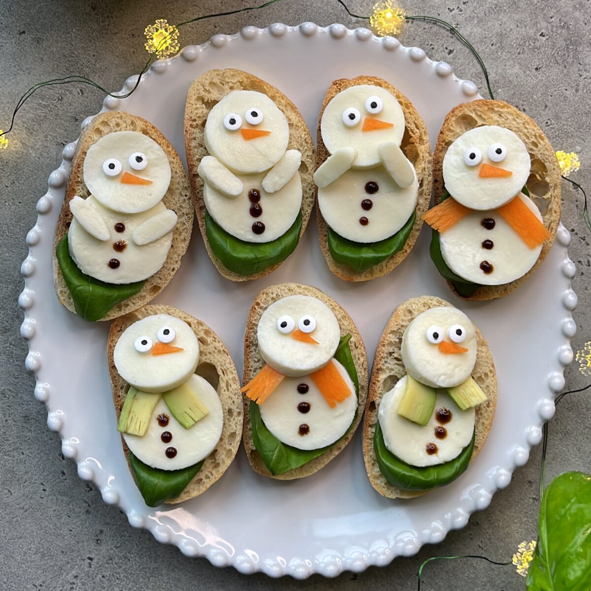 A white plate holds seven bites of bread topped with cheese and vegetables arranged to resemble snowmen, garnished with green leaves and small decorative lights.