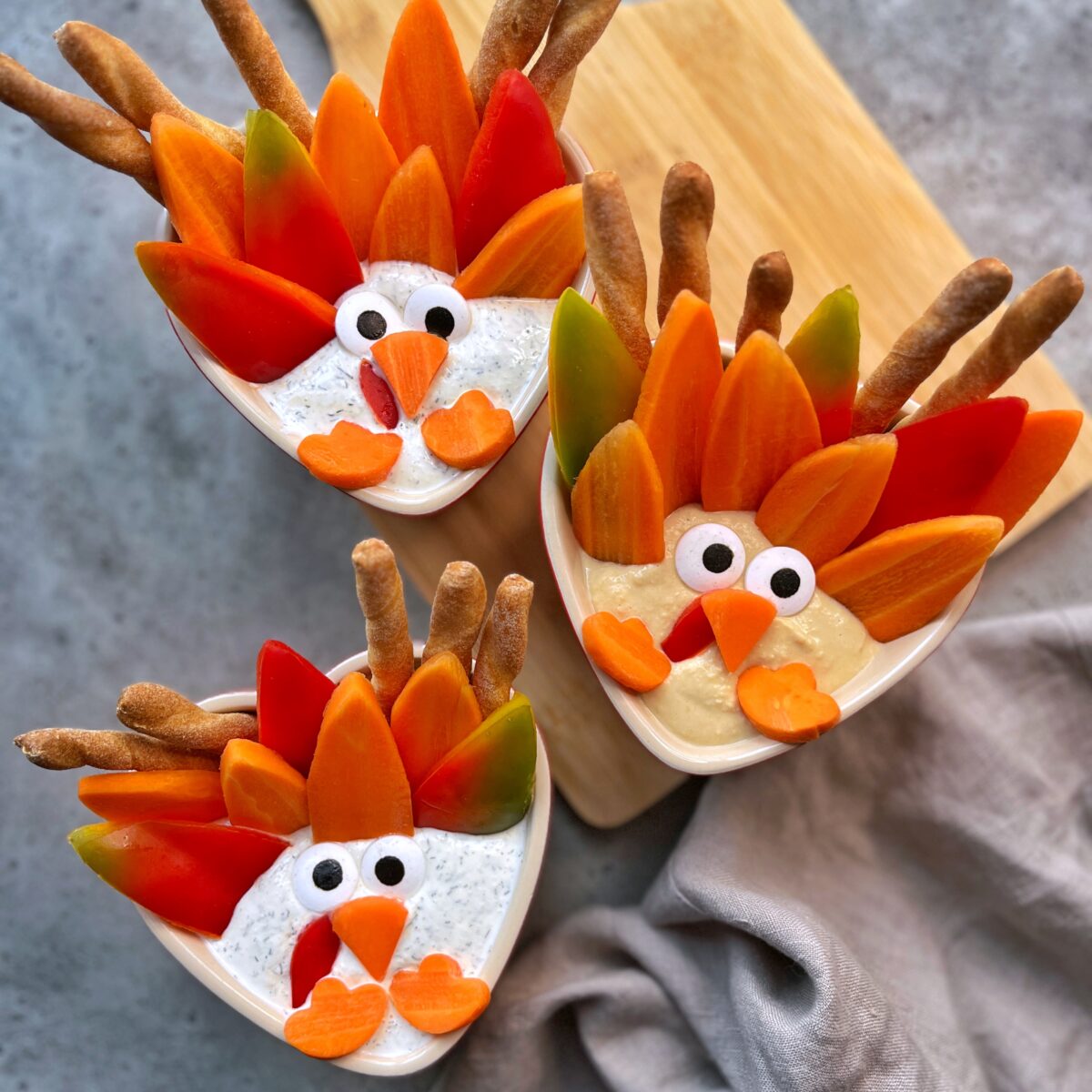 Three bowls of veggie dip decorated to look like turkeys, with carrot and bell pepper feathers, breadstick tails, and faces made from olives and cut vegetables.