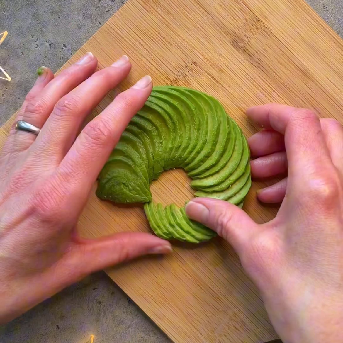 Two hands arranging thinly sliced avocado in a circular pattern on a wooden cutting board.