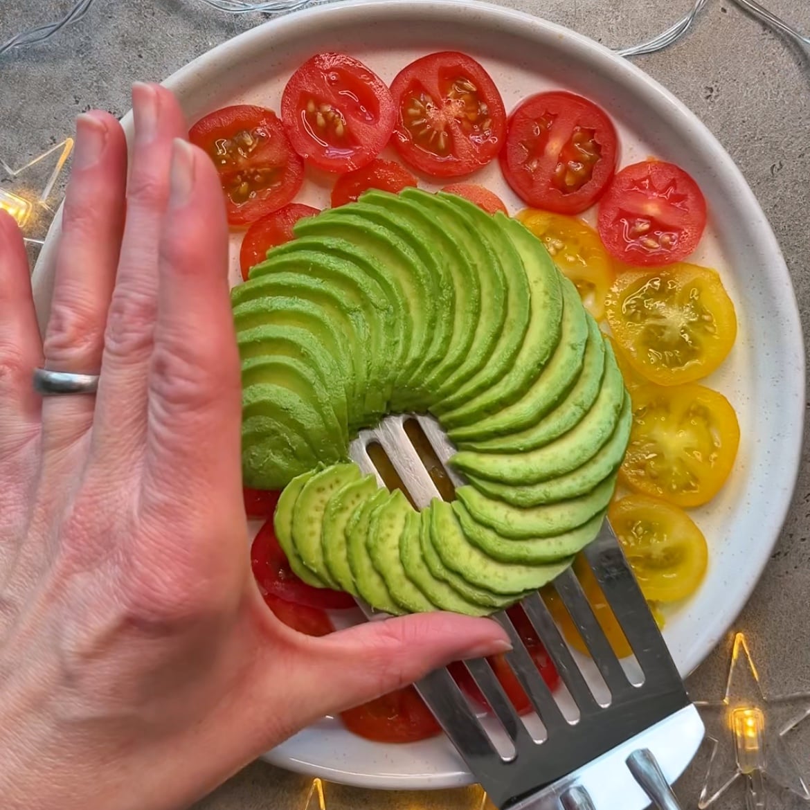 A hand uses a spatula to lift neatly sliced avocado arranged in a spiral on a plate, surrounded by red and yellow tomato slices.