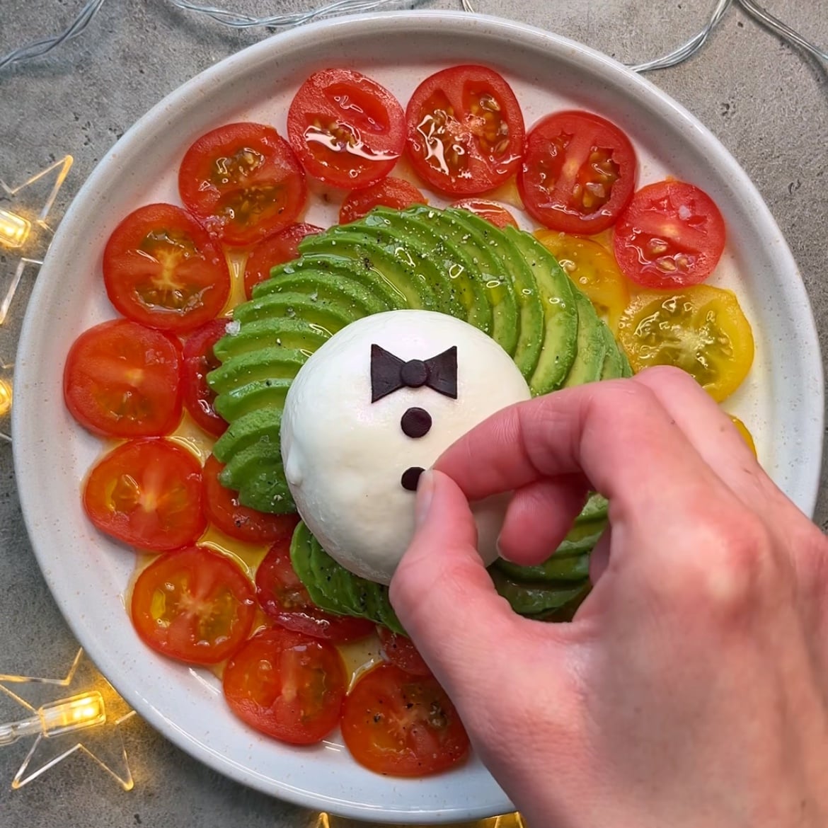 A hand arranges red cabbage 'buttons' on top of a burrata surrounded by avocado and cherry tomatoes.