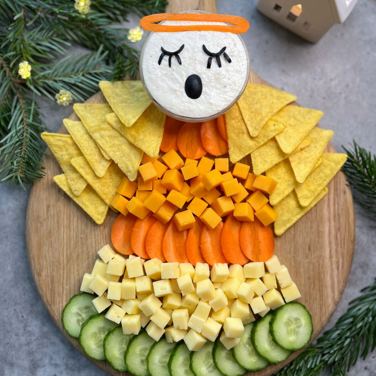 A cheese and vegetable platter arranged to look like an angel, with cucumber, cheese cubes, carrot slices, tortilla chips, and a dip decorated as the angel's face.