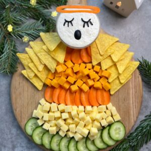 A cheese and vegetable platter arranged to look like an angel, with cucumber, cheese cubes, carrot slices, tortilla chips, and a dip decorated as the angel's face.